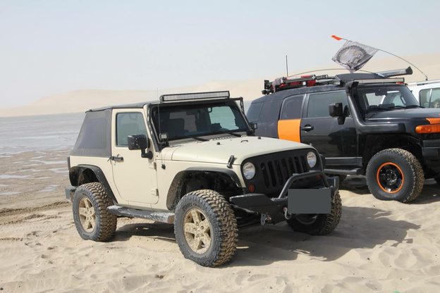 Tan two-door Jeep with a black soft top parked on sand beside a black-and-orange off-road vehicle, with dunes behind them.