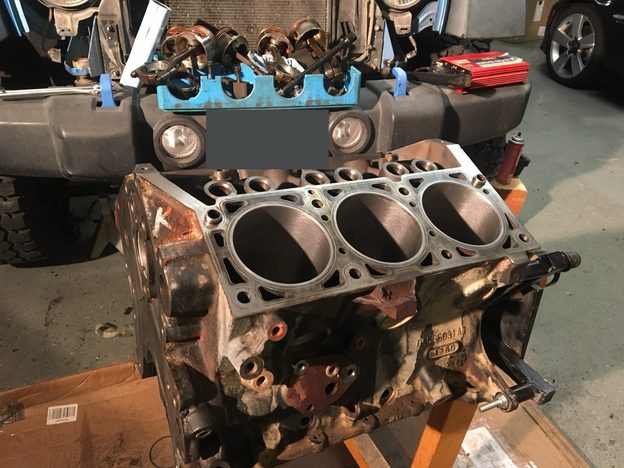 Engine block on an orange stand in front of the blue Jeep, with its cylinder bores open and pistons arranged behind it.
