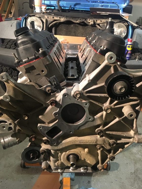 Partly assembled V6 engine on an orange stand, with the open-hood blue Jeep behind it.