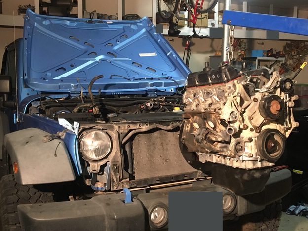 V6 engine suspended from a blue hoist in front of the open-hood blue Jeep in the garage.