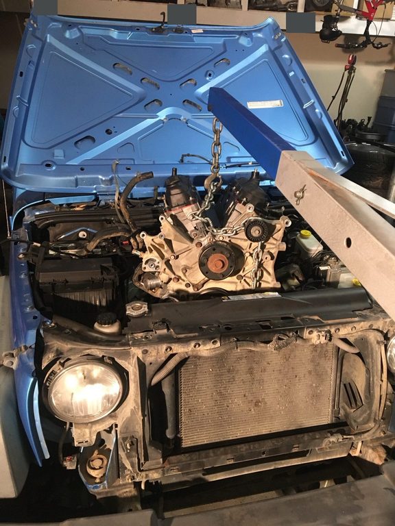 Front view of a V6 suspended over the blue Jeep’s engine bay by a blue hoist.