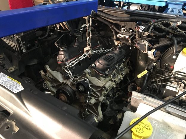 V6 engine in the blue Jeep’s engine bay with hoist chains attached and wiring still disconnected.