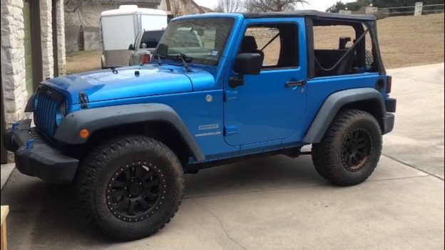 Blue two-door Jeep Wrangler parked on a concrete driveway, with black wheels and the soft-top side windows removed.