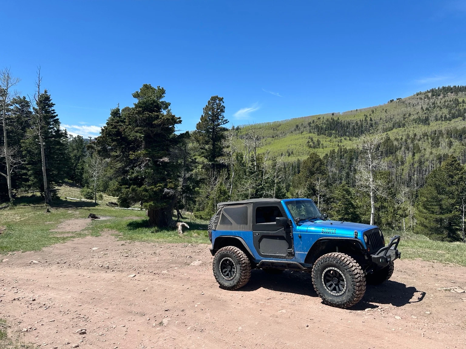 Blue Jeep parked in a dirt clearing with conifers and green hills behind it.