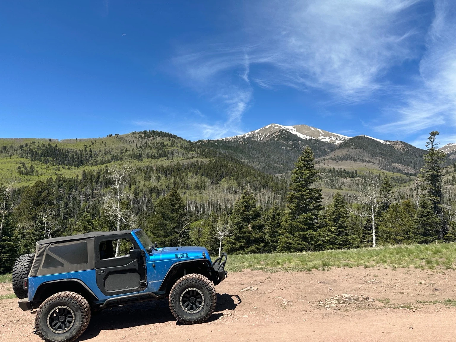 Blue Jeep parked on dirt beneath forested slopes and a snow-streaked mountain.