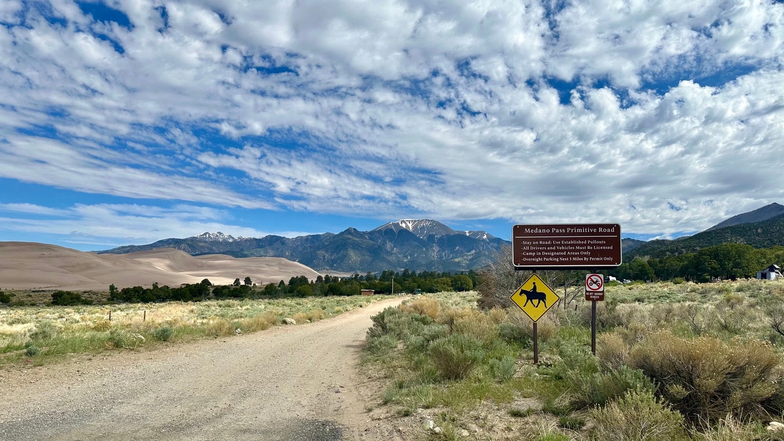 Medano Pass Primitive Road sign beside a dirt road, with sand dunes and mountain peaks in the background.